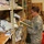 A U.S. Army soldier in ACU (Army Combat Uniform) is conducting an inventory or inspection in a military supply room or storage facility. The soldier is holding a clipboard and appears to be scanning or checking items on metal shelving units. The storage area contains a variety of supplies including boxes, a megaphone, office supplies, and miscellaneous equipment.