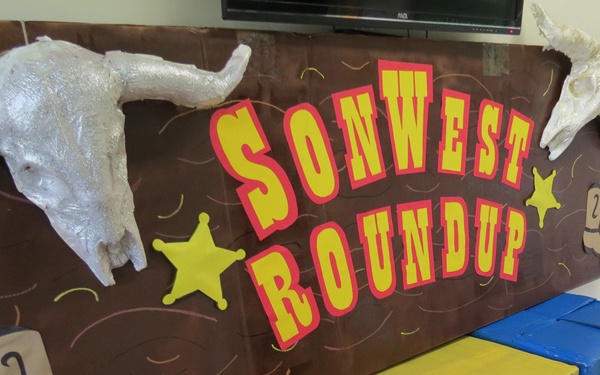 A Western-themed decorative display featuring a large brown banner reading 'SonWest Roundup' with yellow and red lettering. The setup includes prop animal skulls, cowboy boot cutouts, sheriff star decorations, and three colored cardboard boxes labeled 'Animal Feed'. This appears to be a setup for a church vacation bible school or children's event with a Southwest/cowboy theme.
