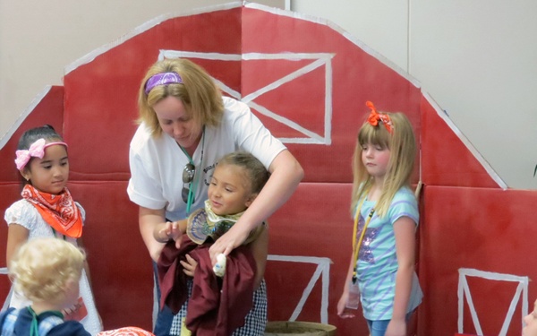 A group of young children are participating in what appears to be a farm-themed school or church play/performance. An adult woman is helping a child with a costume or prop in front of a large painted red barn backdrop. Many children are wearing orange bandanas as part of their costumes.