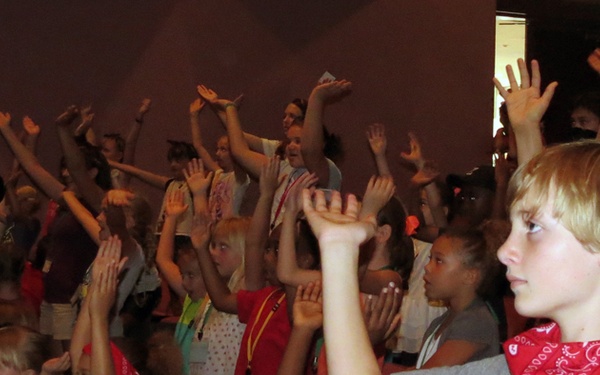 A large group of children and adult supervisors are gathered in what appears to be an auditorium or church hall, with many participants raising their hands in the air. Several children are wearing red bandanas and colored lanyards, suggesting an organized camp or group event. The atmosphere appears energetic and participatory, consistent with a worship service, camp activity, or group game.