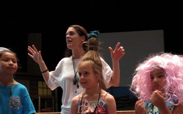 A group of three children and one adult woman appear to be participating in a Vacation Bible School (VBS) activity, likely on a stage or performance area. The adult woman has her hands raised expressively, suggesting she is leading or performing. The children are wearing lanyards and costumes, including a pink wig and a paper plate hat.