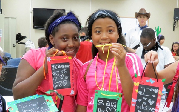 Four young girls pose together at what appears to be a religious summer camp or vacation bible school, each holding handmade craft bags decorated with Christian messages. The girls are smiling and displaying their crafts proudly in an indoor meeting room setting. Adults and other children are visible in the background, some wearing cowboy hats suggesting a Western theme for the event.
