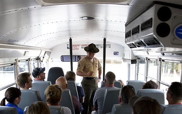 A uniformed military drill instructor or NCO stands at the front of a school bus addressing a group of seated civilians. The bus interior is clearly visible with rows of light blue seats filled with passengers facing forward. The setting appears to be a military base orientation or family day event.