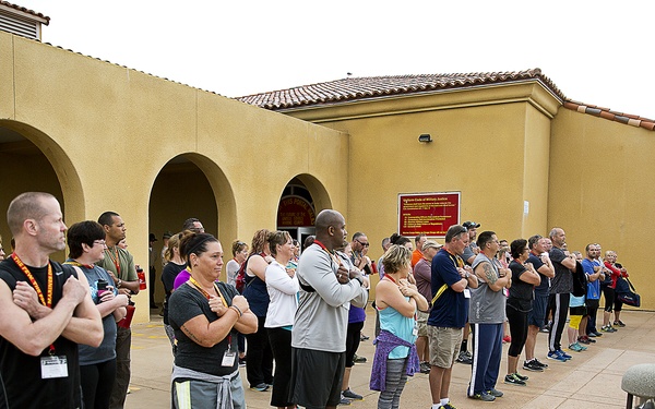 A large group of civilians in casual athletic clothing stands in formation on a paved outdoor area, facing a uniformed military personnel (appearing to be a Marine in khaki uniform). The setting appears to be a military installation, likely MCRD San Diego or a similar Marine Corps base, based on the Spanish Colonial Revival architecture with arched facades and red tile roof. A red sign on the building wall references the Uniform Code of Military Justice (UCMJ), suggesting this may be a civilian orientation or 'family day' type event.