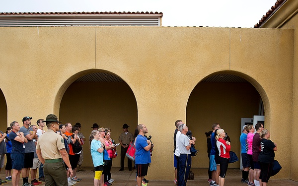 A group of civilians stands in formation on a concrete courtyard featuring painted yellow footprints, overseen by a uniformed Marine Corps drill instructor. The setting appears to be a military base, likely during a family day or civilian orientation program. A red sign referencing the Uniform Code of Military Justice is visible on the wall.