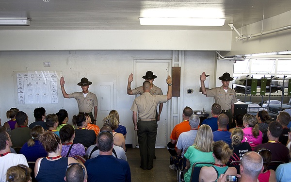 Three uniformed military personnel, appearing to be Marine Corps Drill Instructors wearing campaign covers (drill sergeant hats), stand at the front of a room with their right hands raised. A large civilian audience seated in chairs faces them, suggesting an oath-taking or demonstration ceremony. The setting appears to be a military barracks facility, likely during a family day or public tour event.