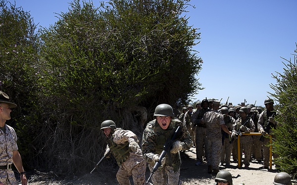 A military training exercise is underway on a sandy outdoor course, with recruits in camouflage uniforms and helmets performing tactical drills including low crawling and rushing movements. A drill instructor wearing a distinctive campaign cover (Smokey Bear hat) observes and oversees the exercise from the left side. A large group of additional recruits in formation can be seen waiting in the background.