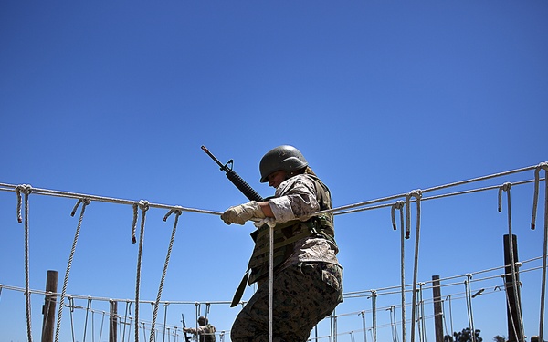 A military recruit in camouflage uniform and helmet navigates a rope obstacle course while carrying a rifle, likely during basic training at a military installation. The wide-angle fisheye lens captures the circular rope bridge structure with wooden posts and gravel/stone borders. Additional personnel in uniform are visible in the background performing similar training activities.