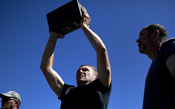 An outdoor physical fitness event or military-style training exercise is taking place under a clear blue sky. A central figure in a black shirt is pressing a dark metal ammunition can overhead with both arms extended, a common military fitness test exercise. Multiple participants are visible in the background, some wearing military camouflage uniforms.