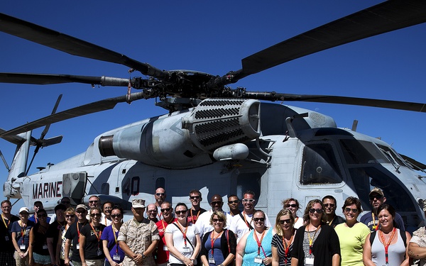 A large group of approximately 30-40 civilians and military personnel pose for a group photo in front of a U.S. Marine Corps helicopter on a tarmac. The helicopter, marked 'MARINE,' appears to be a CH-53 Super Stallion heavy-lift helicopter. The setting appears to be a military air station during what looks like a community outreach or familiarization tour event.