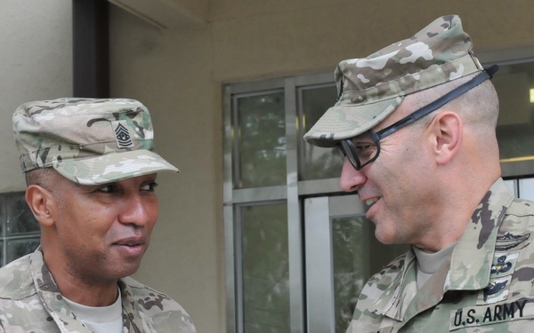 Two U.S. Army soldiers in Operational Camouflage Pattern (OCP) uniforms are engaged in a handshake outside a building. The soldier on the left has the name tape 'THOMAS' and wears a '1st Infantry Division' patch, while the soldier on the right has the name tape 'SPANO' and wears glasses. Both appear to be senior Non-Commissioned Officers based on their rank insignia.