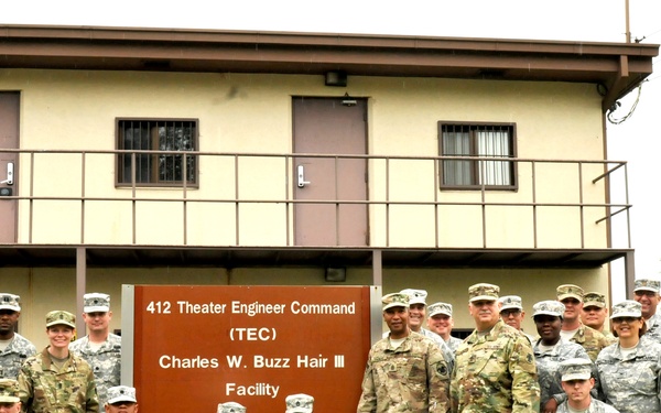 A group of approximately 25 U.S. Army soldiers in military uniforms pose for a group photograph in front of a sign identifying the '412 Theater Engineer Command (TEC) Charles W. Buzz Hair III Facility.' The soldiers are arranged in multiple rows, with some kneeling in front and others standing behind, in front of a two-story beige military building. The image appears to be an official unit or command photo.