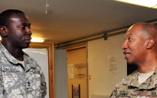 Two U.S. Army soldiers are shaking hands in what appears to be an indoor military facility. The soldier on the left wears ACU (Army Combat Uniform) digital camouflage with the name tape 'BURGESS' and a III Corps patch, while the soldier on the right wears MultiCam/OCP uniform with the name tape 'THOMAS' and a Sergeant Major rank insignia. The interaction appears formal and ceremonial in nature.