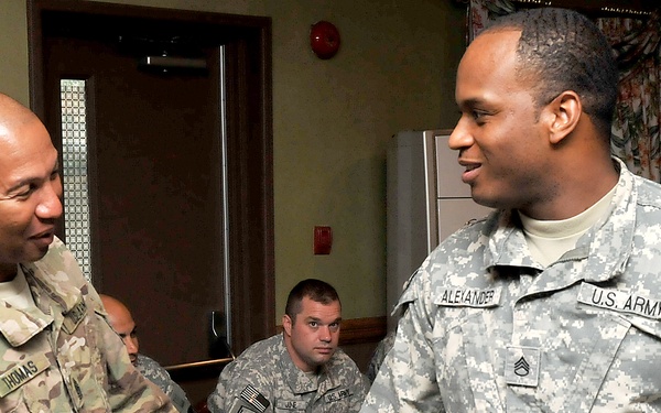 Two U.S. Army soldiers are shaking hands in what appears to be an indoor dining or meeting facility. The soldier on the left wears a MultiCam (OCP) uniform with the name tape 'THOMAS' and a 1st Infantry Division patch, while the soldier on the right wears an ACU (Army Combat Uniform) with the name tape 'ALEXANDER' and a Staff Sergeant rank insignia. Several other soldiers are seated in the background observing the interaction.