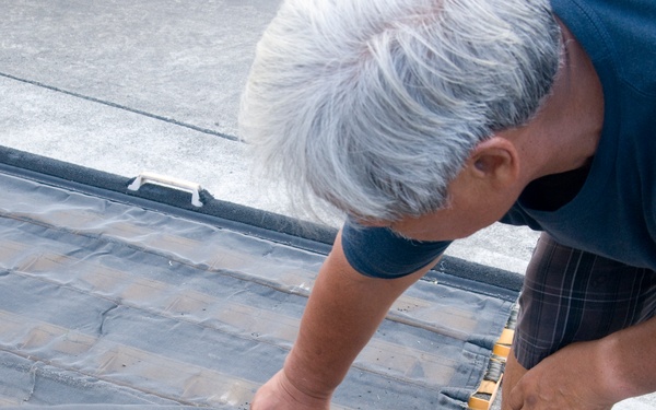An older man with white/gray hair is bending over to inspect or handle a dark rubber or membrane material on what appears to be a flat surface with yellow structural framing. The material appears worn or damaged, with visible tears or deterioration. The scene suggests a maintenance or inspection activity, possibly related to a loading dock, ramp, or flat roof.