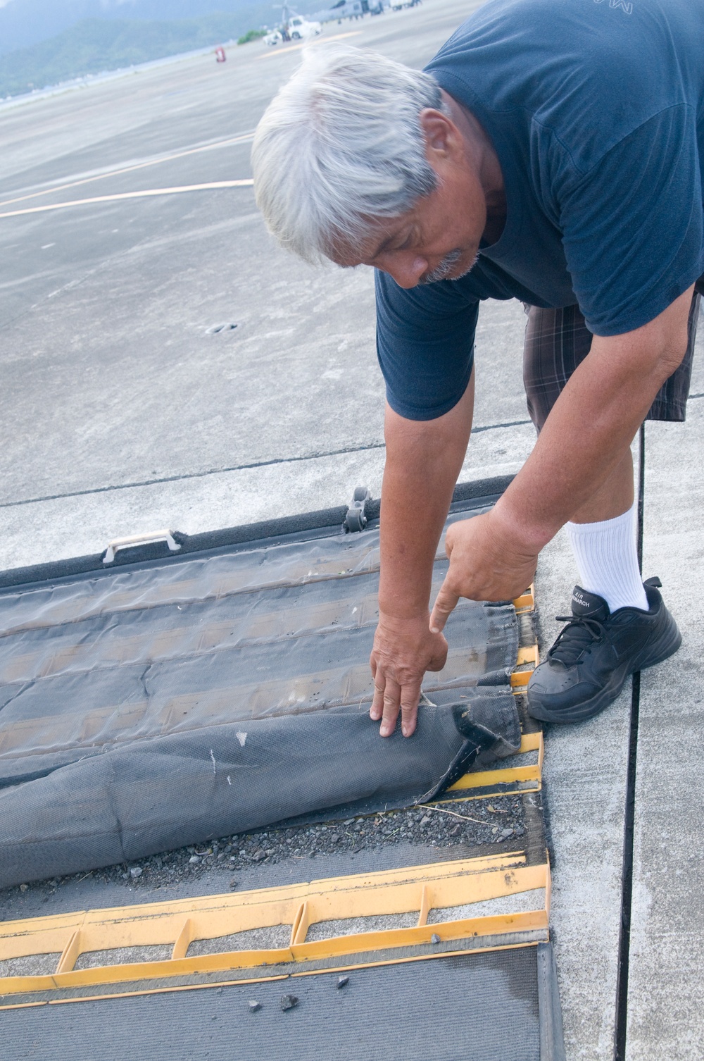 A middle-aged man with white/gray hair is bending over and inspecting or adjusting a black rubber or fabric seal/gasket along what appears to be an expansion joint or threshold on a concrete surface, likely an airport tarmac or apron. He is pointing at or pressing down on the material, suggesting maintenance or inspection work. The background shows an open airfield with distant vehicles and green hills.