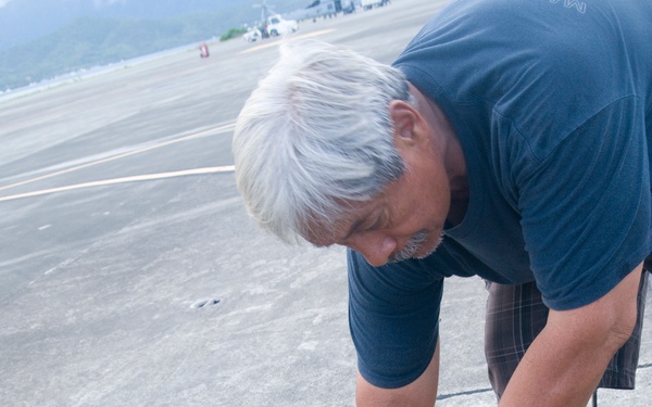 A middle-aged man with white/gray hair is bending over and inspecting or adjusting a black rubber or fabric seal/gasket along what appears to be an expansion joint or threshold on a concrete surface, likely an airport tarmac or apron. He is pointing at or pressing down on the material, suggesting maintenance or inspection work. The background shows an open airfield with distant vehicles and green hills.