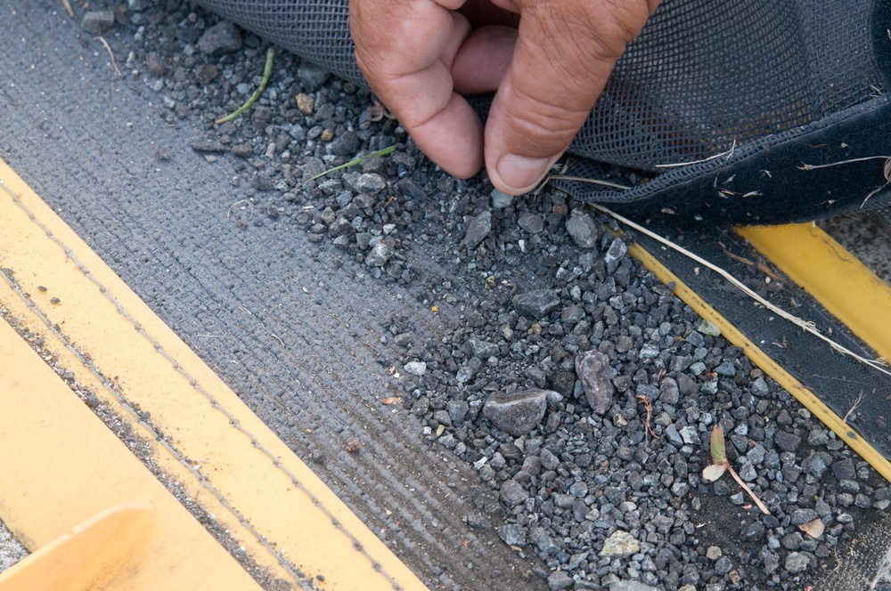 A close-up image shows a person's hand lifting or pulling back a dark mesh/fabric material from a surface covered with gravel and small stones. The surface appears to be a textured platform or walkway with yellow safety markings or edging visible on the left and right sides. The scene suggests maintenance, inspection, or installation work involving drainage or erosion control materials.