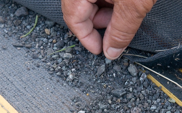 A close-up image shows a person's hand lifting or pulling back a dark mesh/fabric material from a surface covered with gravel and small stones. The surface appears to be a textured platform or walkway with yellow safety markings or edging visible on the left and right sides. The scene suggests maintenance, inspection, or installation work involving drainage or erosion control materials.