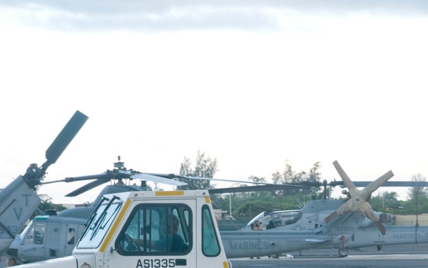 The image shows a military flight line with a white aircraft ground support tug (TUG model AS1335, GPMK) in the foreground on a concrete apron. Behind the tug, multiple U.S. Marines helicopters are parked, identifiable by 'MARINES' markings on their fuselages. The setting appears to be a tropical military airfield based on the vegetation visible in the background.