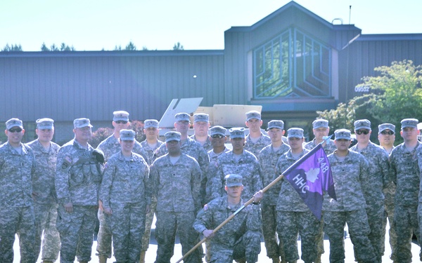 A group of approximately 20 U.S. Army soldiers in ACU (Army Combat Uniform) camouflage are posed for a formal unit photograph. One soldier kneels in the front center holding a purple guidon flag with 'HHC' and what appears to be '148' visible. The group is positioned on a paved area flanked by military Humvee vehicles with a building and trees visible in the background.