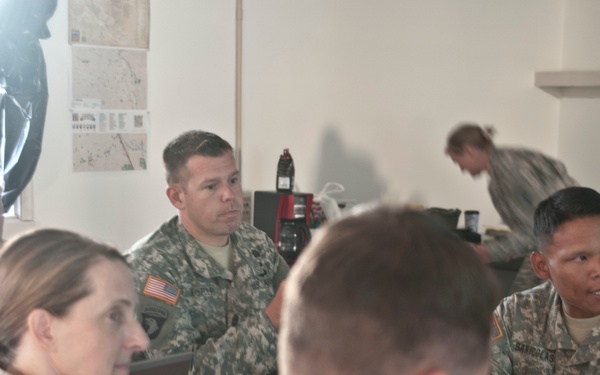 A group of U.S. Army soldiers in ACU (Army Combat Uniform) camouflage are gathered around a wooden table in what appears to be a briefing or planning session. Multiple soldiers are engaged in discussion, with maps and documents visible on the wall behind them. The setting appears to be a forward operating base or military operations center.