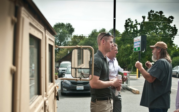 Four individuals are engaged in conversation in a parking lot beside a tan/beige military-style HMMWV (Humvee) vehicle. Two men on the left appear to be in professional or semi-professional attire, while the two individuals on the right are dressed casually. The setting appears to be a commercial area with parked cars, trees, and signage visible in the background under an overcast sky.