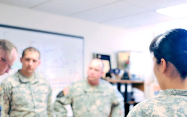 The image shows a briefing or meeting involving military personnel in ACU (Army Combat Uniform) camouflage and a civilian law enforcement officer in a dark uniform. The setting appears to be an office or operations room with a whiteboard visible in the background. A female soldier in the foreground displays a patch consistent with US Army Cyber Command or a similar unit, along with an Airborne tab.