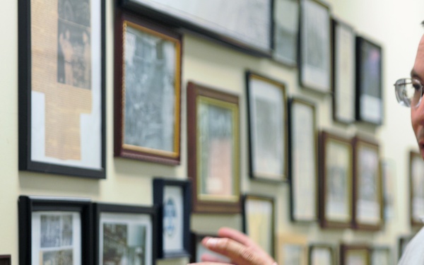 A middle-aged man wearing glasses and a polo shirt with a logo patch is gesturing with his hands while speaking, standing in front of a wall densely covered with framed photographs and documents. The setting appears to be a museum, archive, or historical display room. He appears to be giving a tour or explanation of the historical materials on the wall.
