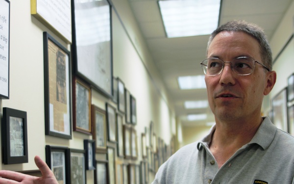 A middle-aged man wearing glasses and a gray polo shirt with an 'Airborne' military patch is standing in a hallway lined with framed photographs, documents, and memorabilia. He appears to be giving a tour or presentation, gesturing toward the wall displays with his right hand. The setting appears to be a military museum, archive, or historical display corridor.