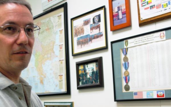 A middle-aged man wearing glasses and a gray polo shirt is pointing to a framed photograph on a wall covered with military memorabilia, certificates, maps, and awards. The wall displays numerous framed items including medals, unit citations, certificates of achievement, photographs of military personnel, and what appears to be a deployment roster with small flags. The setting appears to be a military office or personal display room commemorating service history.