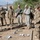A group of approximately eight to nine military personnel in camouflage uniforms are gathered outdoors on a dirt surface, appearing to conduct a training exercise or briefing. Some soldiers are taking notes on clipboards while one examines objects on the ground using a stick or rod. The setting appears to be a military installation with buildings and utility poles visible in the background.