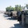 A convoy of military HMMWV (Humvee) vehicles is staged along a gravel road in an arid, desert-like environment. A soldier in ACU (Army Combat Uniform) camouflage pattern is standing at the open door of the nearest Humvee. The setting appears to be a small rural town or base area with utility poles, trees, and low hills visible in the background.