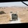 The image is taken from inside a military vehicle looking through the windshield at a convoy of olive-drab military trucks traveling along a narrow gravel road through dry, golden rolling hills. At least two military trucks are visible ahead, with additional vehicles further in the distance. The terrain appears arid with sparse oak trees and dry grass, consistent with a California training range environment.