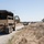 A convoy of military tactical vehicles is traveling along a gravel road through dry, open grassland terrain. The vehicles are painted in desert tan/sand color scheme typical of U.S. military equipment. The landscape appears arid with dried grass and scattered oak trees, consistent with central California terrain.
