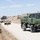 A U.S. military convoy is halted on a dirt road in a dry, hilly terrain consistent with a training environment in California. The convoy consists of at least one LMTV (Light Medium Tactical Vehicle) and multiple HMMWVs (Humvees), with soldiers dismounted and taking up security positions along the roadside. The scene appears to be a convoy security drill or field training exercise (FTX).