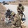 The image shows U.S. Army soldiers conducting a combat casualty care training exercise along a rural dirt road. Two soldiers are administering simulated medical treatment to a fellow soldier lying on the ground, while a third armed soldier stands guard. A military Humvee is visible in the background on the road.