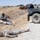 The image depicts what appears to be a military training exercise in an arid, open field environment. Two soldiers are lying on the ground simulating casualties while a third soldier kneels nearby, likely acting as a combat medic providing simulated treatment. A military HMMWV (Humvee) is parked on a gravel road to the right, with an additional soldier positioned near or on the vehicle.