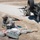 The image depicts what appears to be a military training exercise involving three soldiers in U.S. Army camouflage uniforms. One soldier is providing simulated medical aid to a downed soldier on the ground, while a second soldier takes a defensive firing position behind a military Humvee. Medical supplies are visible scattered near the casualty, indicating a combat casualty care drill.