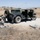 The image depicts a military training exercise in an arid, open terrain environment. Several soldiers in U.S. Army combat uniforms are positioned around a military HMMWV (Humvee), with one soldier aiming a rifle, one crouching with equipment, and one lying prone on the ground. The setting appears to be a training range, likely at a U.S. military installation based on the landscape and equipment.
