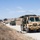 A military convoy of tactical vehicles is halted on a dirt road in an arid, hilly training environment. The lead vehicle, a tan HEMTT (Heavy Expanded Mobility Tactical Truck) marked 'A-576' and '40CAB/640', is stopped with soldiers taking up positions along the roadside. The scene appears to depict a training exercise, likely at a U.S. Army installation such as Fort Hunter Liggett or Camp Roberts in California.