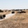 The image shows a dry, open grassland terrain with a gravel road running through it. Two military Humvees are parked along the road, and a group of personnel are gathered near what appears to be a heavily rusted and destroyed armored vehicle or tank hulk on the right side of the road. The setting appears to be a military training range or installation, likely in a semi-arid region of the western United States.