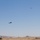 Two military helicopters are flying over an open, arid grassland terrain with rolling hills in the background. A military ground vehicle, likely a HMMWV (Humvee), is partially visible in the lower right corner. The scene suggests a military training exercise or operational movement in a semi-arid landscape.