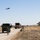 A military field exercise is taking place on a dirt road in an arid, open landscape. Two military vehicles are stopped on the road with soldiers positioned around them, while a helicopter flies overhead. The scene appears to be a training operation or convoy security drill.
