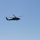 A military helicopter is captured in flight against a clear, cloudless blue sky. The aircraft appears to be a dark olive/green colored utility helicopter, consistent with a UH-60 Black Hawk type, photographed from below at a moderate distance. The rotor blades show motion blur, indicating active flight.