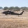 A U.S. Army UH-60 Black Hawk helicopter configured as a MEDEVAC (medical evacuation) aircraft is hovering low over dry, golden grassland. The helicopter displays a red cross emblem on its fuselage, indicating its medical mission role. The setting appears to be a training area in a semi-arid environment, likely in California based on the oak-covered hills in the background.