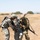 Three military personnel are visible in an open, dry grassland terrain. Two soldiers are assisting a third who appears to have a white bandage wrapped around their head, suggesting a simulated or actual casualty scenario. The scene appears consistent with a field training exercise or combat casualty care drill.