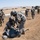 The image depicts what appears to be a U.S. Army field training exercise in an arid, open landscape. Two soldiers wearing helmets are performing a casualty care drill on a simulated downed soldier on the ground, while other soldiers observe or stand nearby. Military vehicles are visible in the background along a dirt road.