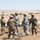 A group of U.S. military personnel are conducting what appears to be a field medical evacuation exercise or training scenario in an arid, open landscape. Several soldiers in combat uniforms and helmets are handling a military stretcher/litter, with at least one person lying on the ground simulating a casualty. The setting appears to be a military training installation, likely in California based on the terrain.