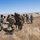 A group of military personnel in camouflage uniforms are conducting what appears to be a casualty evacuation (CASEVAC) training exercise in an open, arid field. Soldiers are carrying a simulated casualty on a litter/stretcher while others attend to additional casualties on the ground nearby. The setting, terrain, and organized nature of the activity suggest a field training exercise at a military installation.